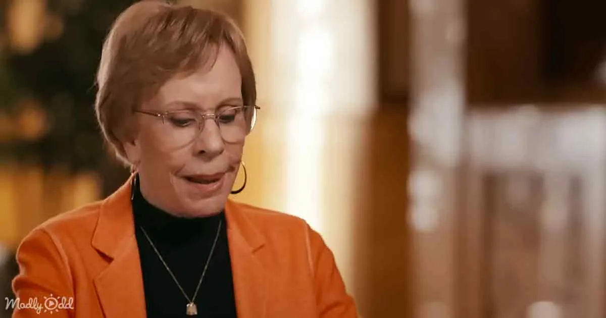 A woman in an orange blazer and glasses is looking down, possibly in thought or sadness.