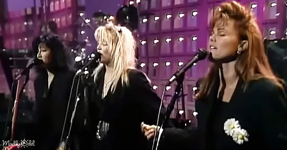 Three women sing into microphones on stage, wearing black, with a cityscape backdrop.