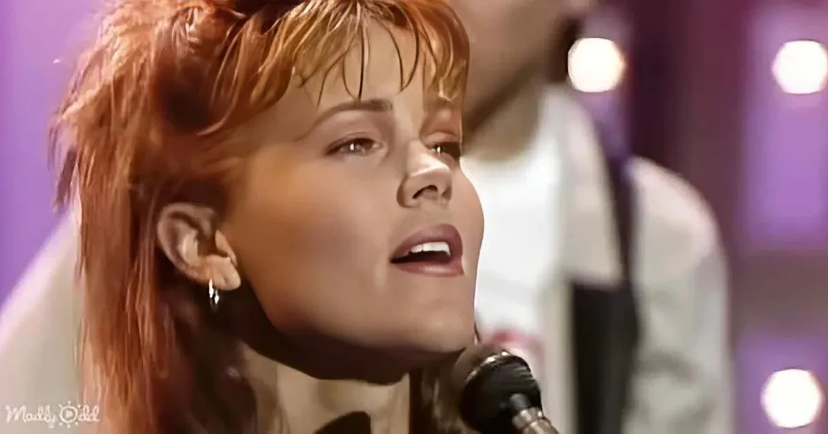 A woman with red hair and a microphone, wearing a white outfit, singing on stage.