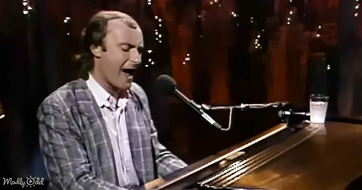 A man in a plaid shirt sings passionately at a piano, with a microphone and glass of water nearby.