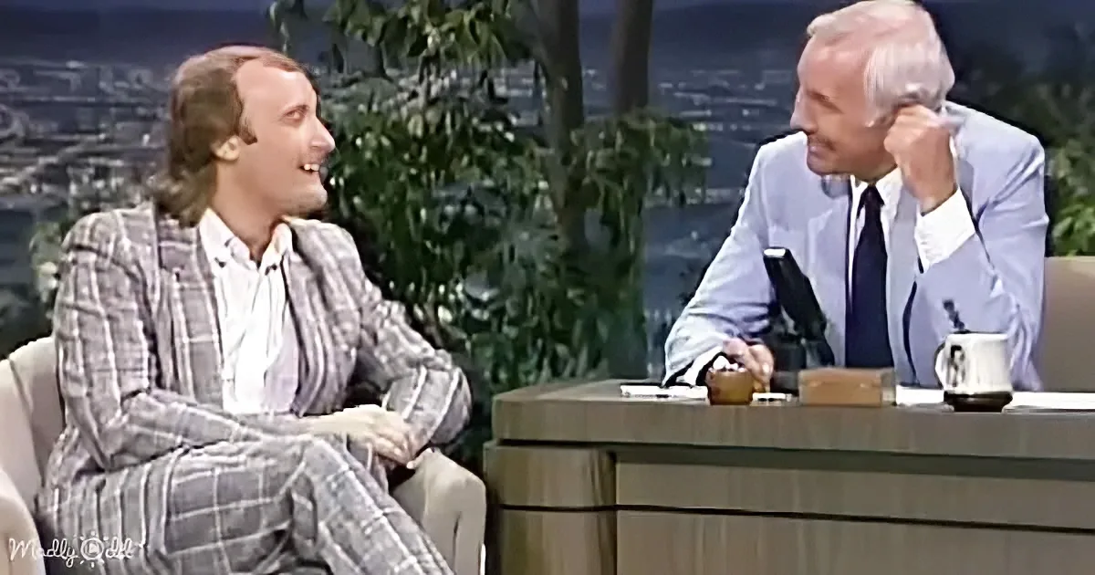 A man in a plaid suit sits on a couch, smiling, while another man in a suit leans forward, appearing to listen intently.