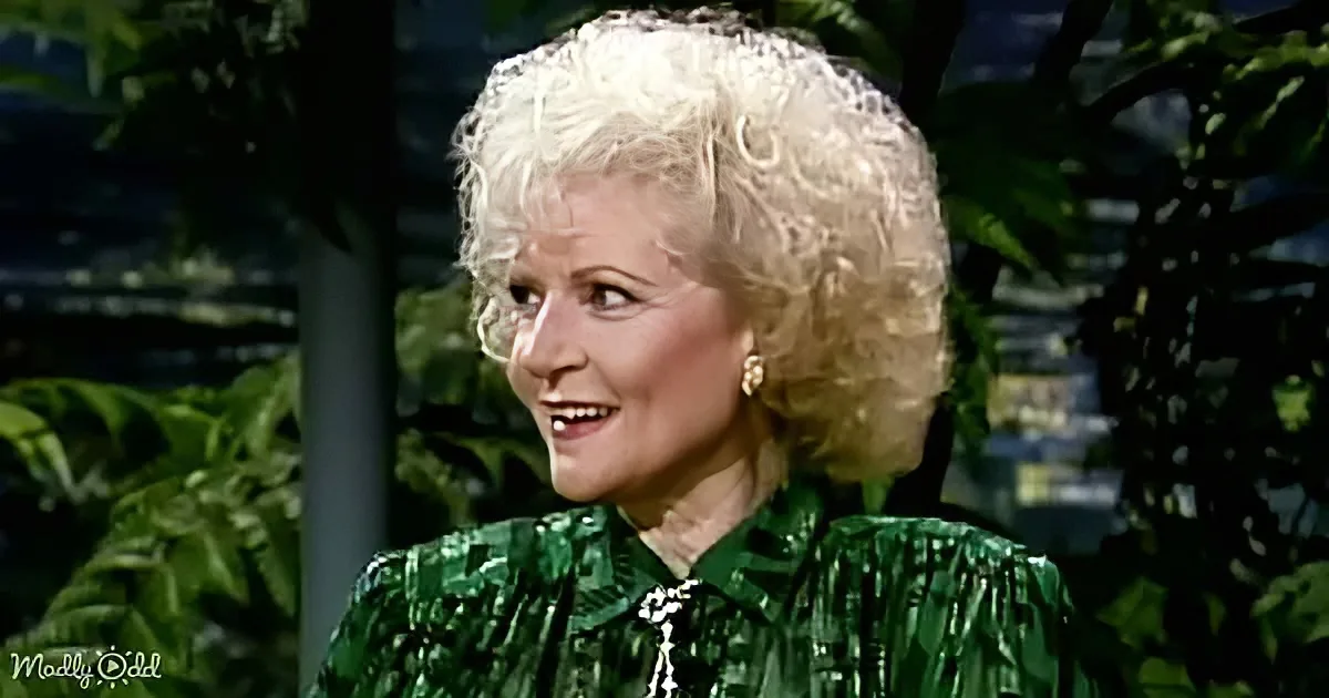 A woman with blonde, curly hair in a green, fringed top smiles, surrounded by greenery.