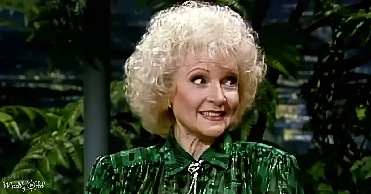 A woman with curly white hair, wearing a green sequined top, smiles while looking to the side.