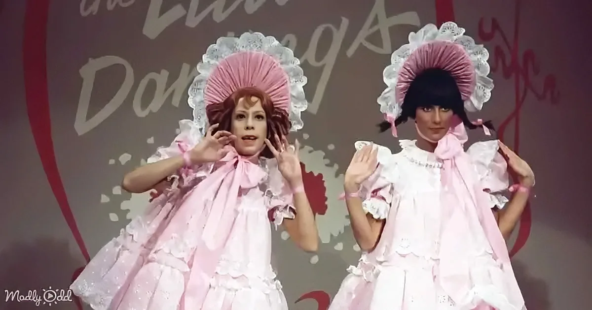 Two individuals in elaborate pink dresses with large hats, posing on a stage.