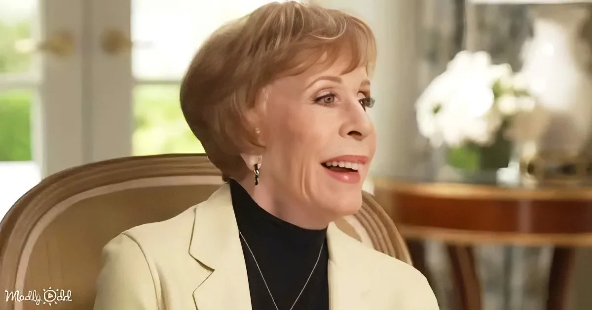 A woman with short, light brown hair, wearing a white blazer and black turtleneck, sits in a chair, smiling.