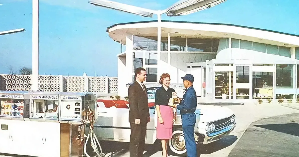 A man in a suit and a woman in a pink skirt stand at a gas pump, talking to a gas station attendant.