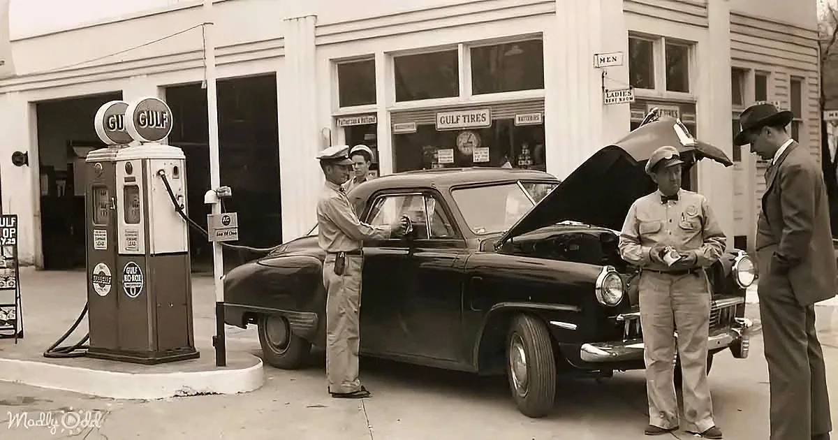 A man in uniform stands by a vintage car at a Gulf gas station, interacting with another worker.