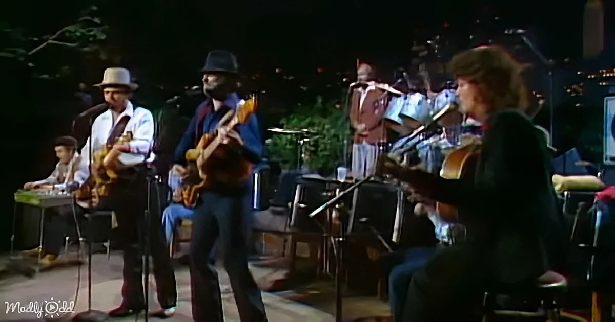 A man in a hat plays guitar while a band performs on stage with microphones and instruments.