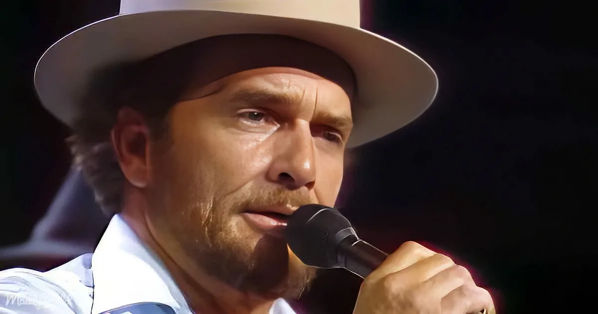 A man in a white hat and shirt speaks into a microphone, holding it with his right hand.