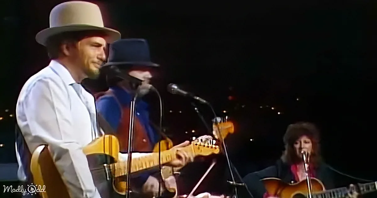 A man in a white shirt and hat plays guitar on stage with a band.