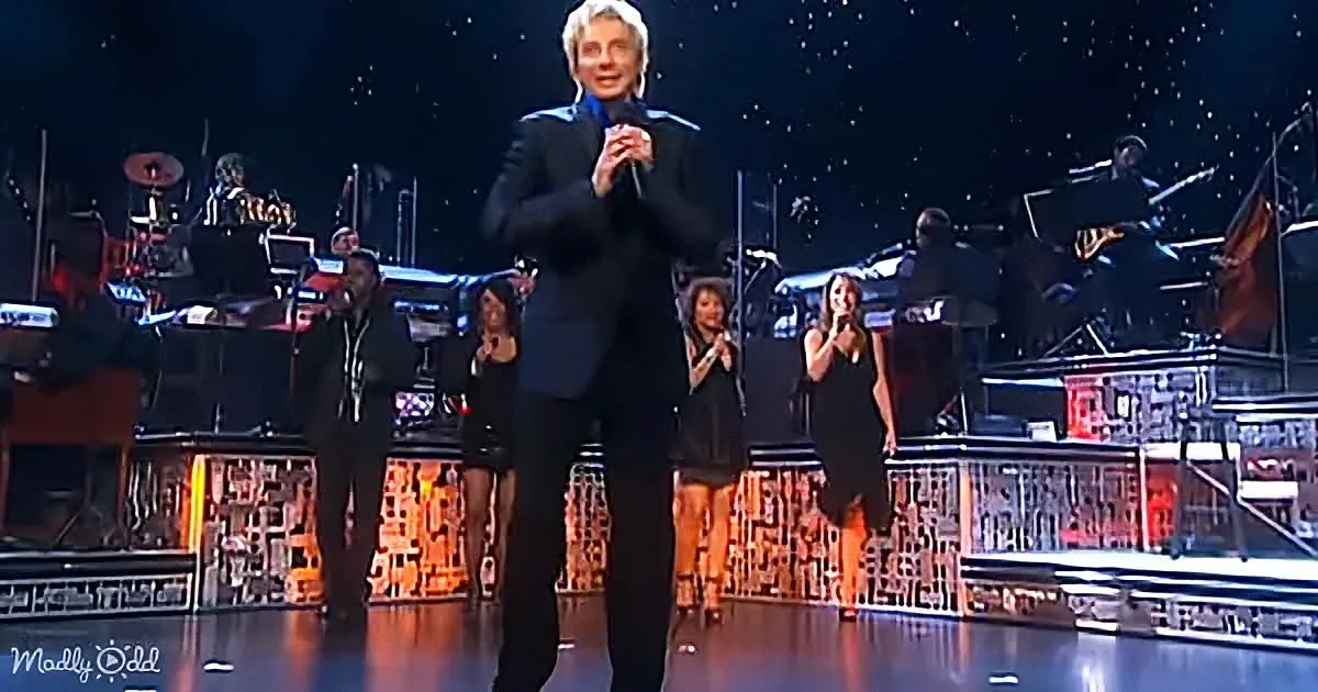 A man in a black suit stands center stage, clapping, with a band and dancers behind him.