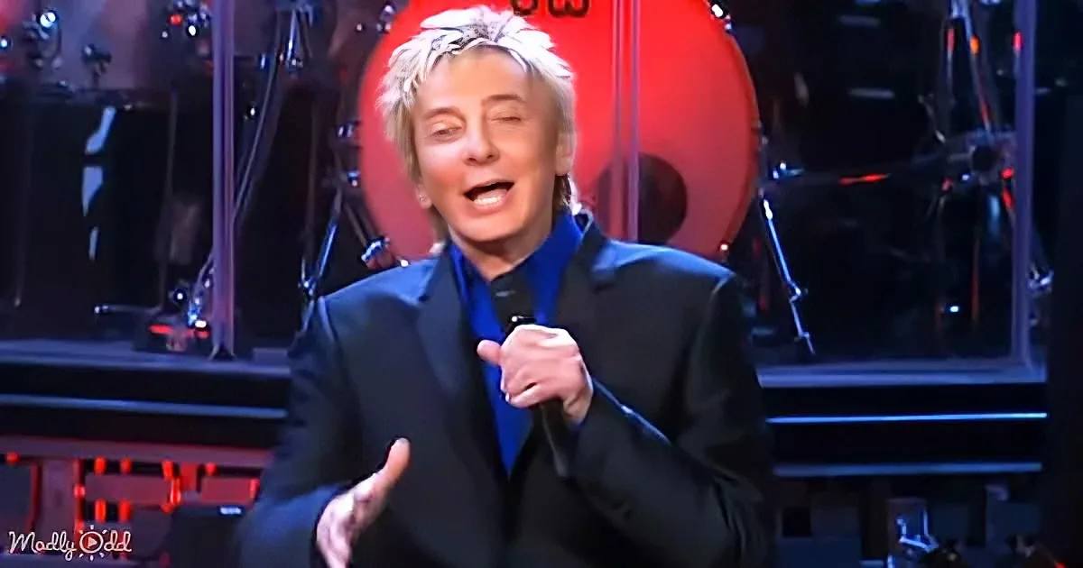 A person in a black suit and blue shirt sings into a microphone on stage with a red drum in the background.