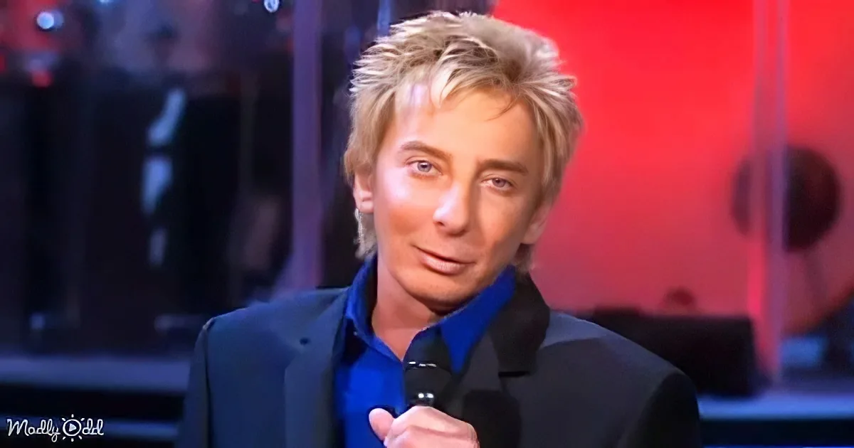A person with blonde hair, wearing a blue shirt and black jacket, holding a microphone, standing in front of a red...