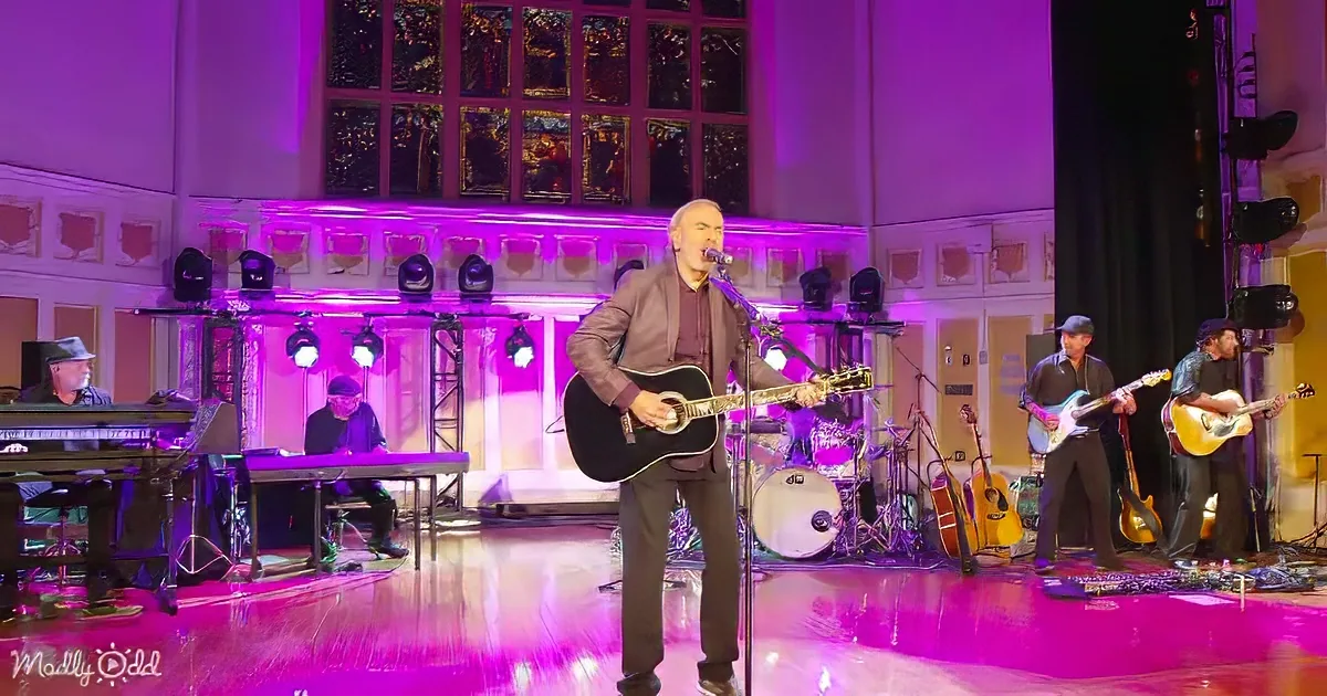 A man plays guitar and sings on stage with a band, illuminated by purple lights.