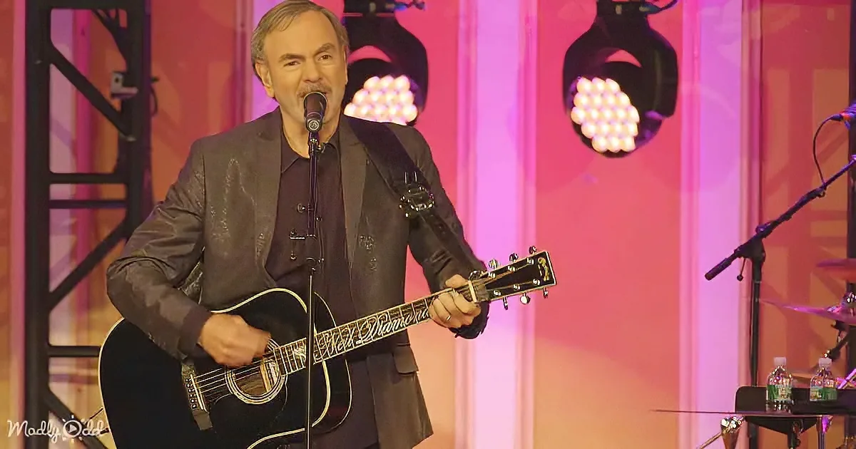 Neil Diamond performs on stage, holding guitar, singing into microphone, with pink and purple lighting.