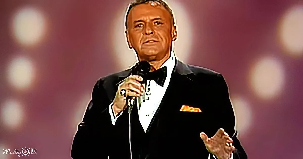 A man in a tuxedo holding a microphone, gesturing, with a blurred background of lights.