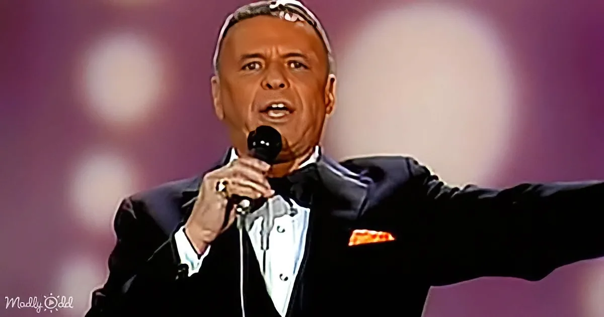 A man in a tuxedo sings into a microphone, gesturing with his right arm extended.