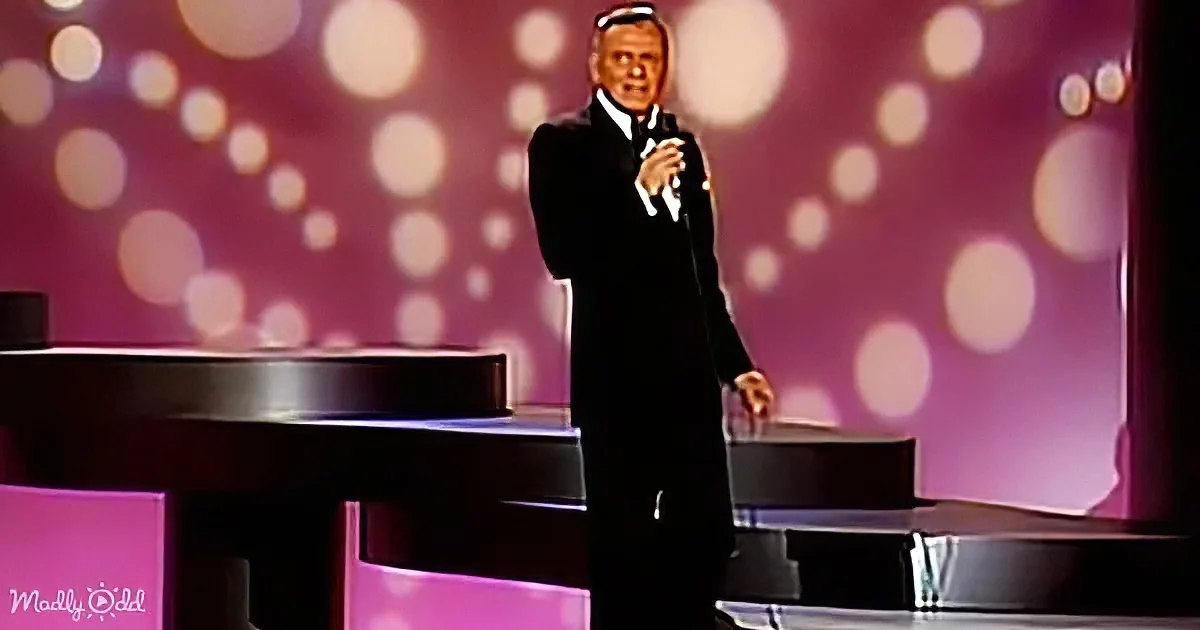 A man in a tuxedo stands on stage with a microphone, performing in front of a piano and a pink, bokeh-lit backdrop.
