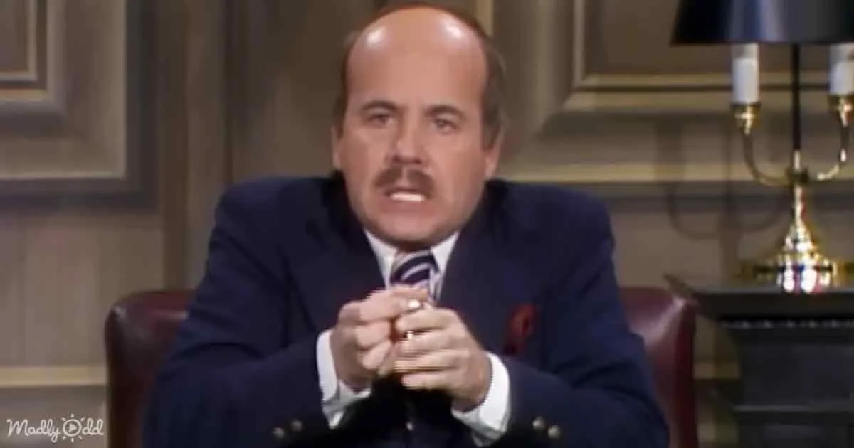 A man in a suit sits with clasped hands, wearing a tie, in a formal setting.