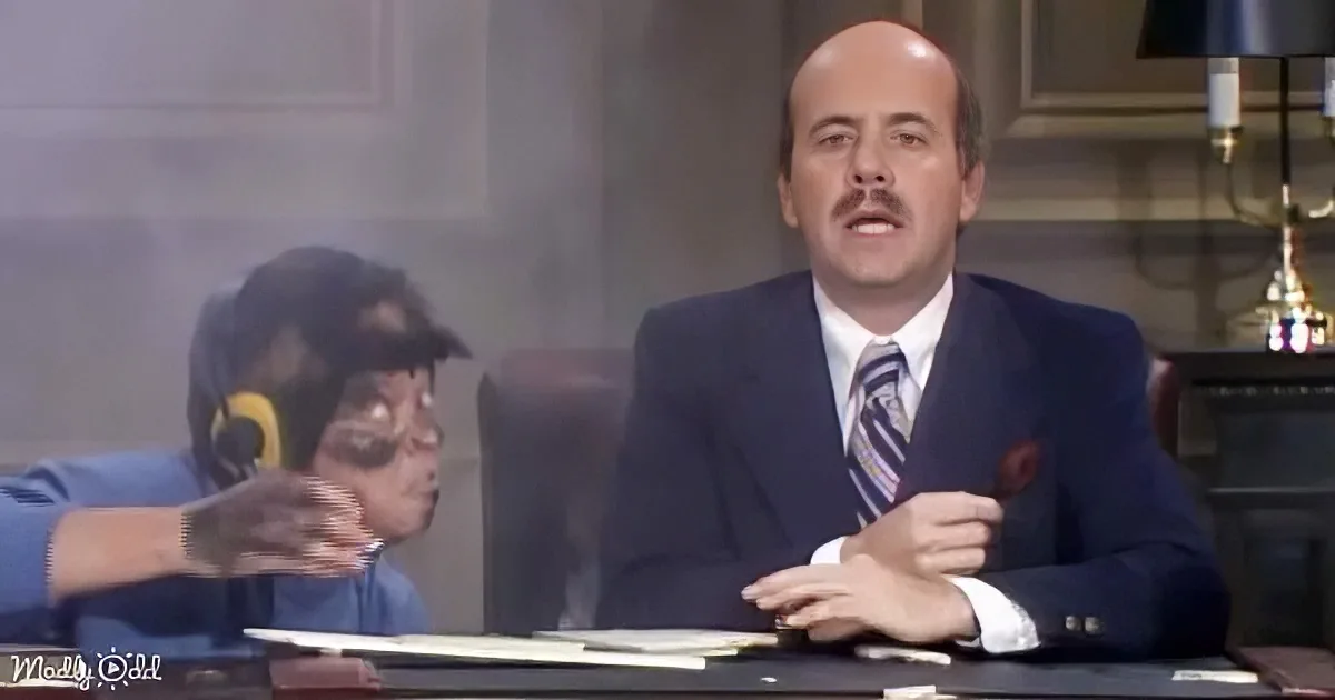 A man in a suit sits at a desk, gesturing with his hands, while another person in a headset leans forward.