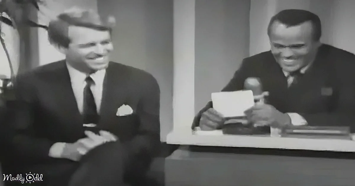 Two men in suits sit at a desk, one holding a microphone, both smiling.