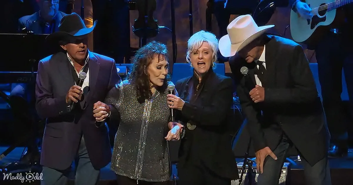 Four performers in cowboy hats sing on stage, holding microphones, with a band in the background.