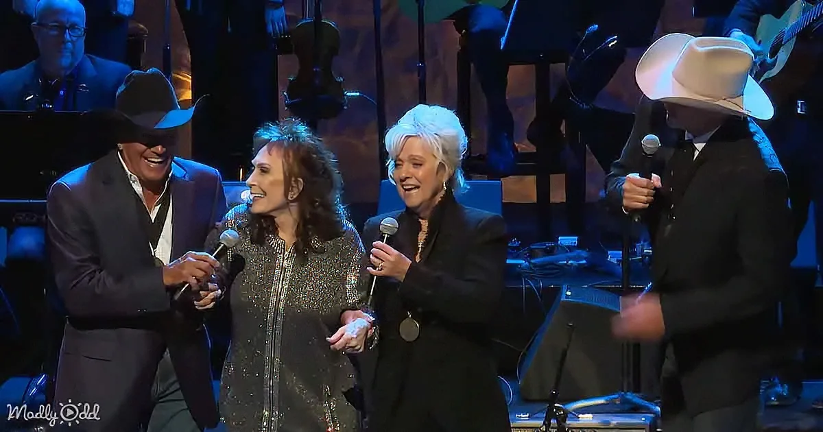 A group of people, including a man in a cowboy hat, are on stage holding microphones, smiling and laughing.