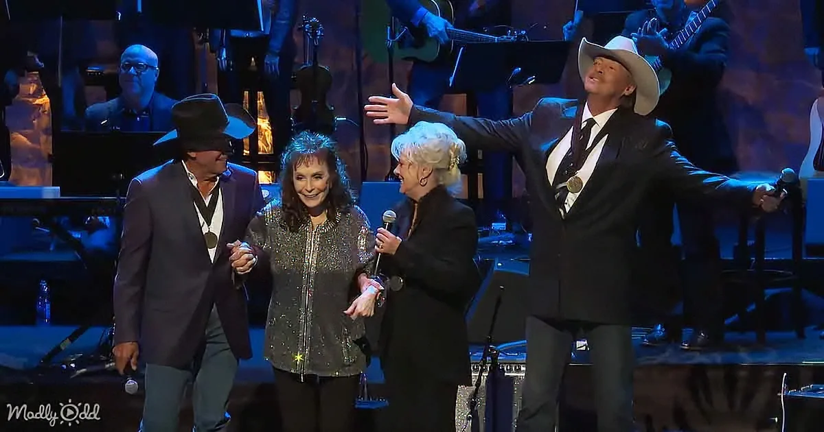 A man in a cowboy hat and suit is raising his hand, smiling, with a woman in a glittery jacket and another in a black...