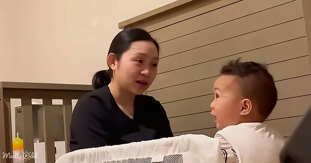 A woman in a black shirt interacts with a child in a white shirt near a crib, with a bottle of orange juice visible.