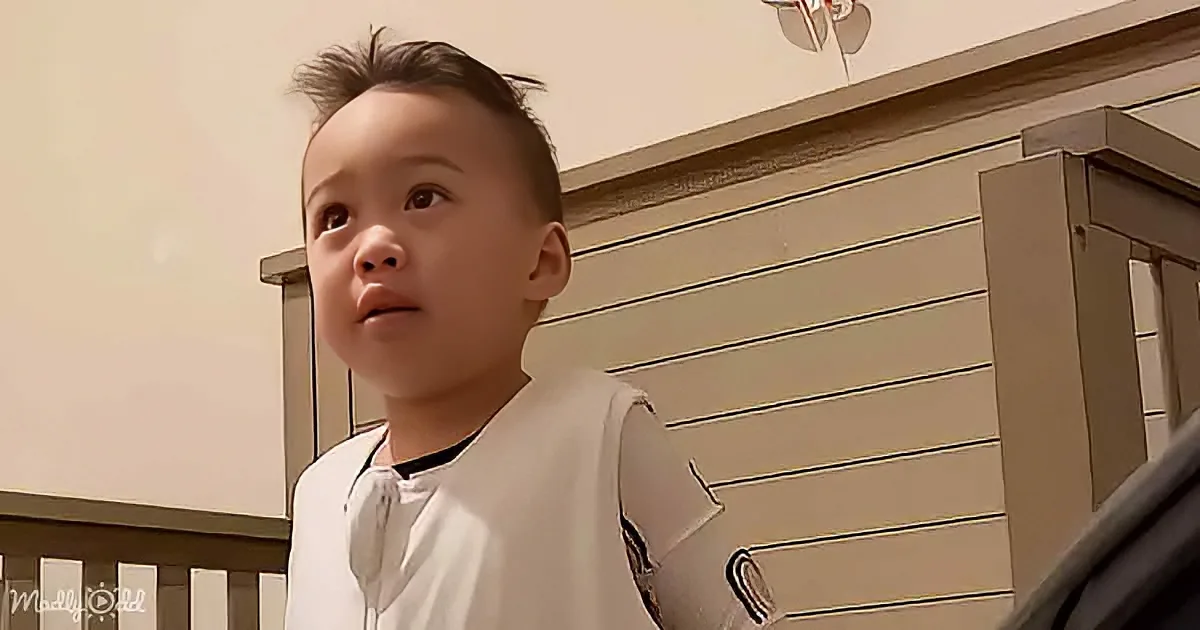 A young child with short hair and a white shirt stands indoors near a staircase.