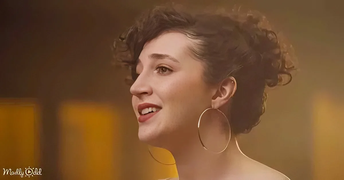 A woman with curly hair, wearing a yellow top and large hoop earrings, smiles softly.
