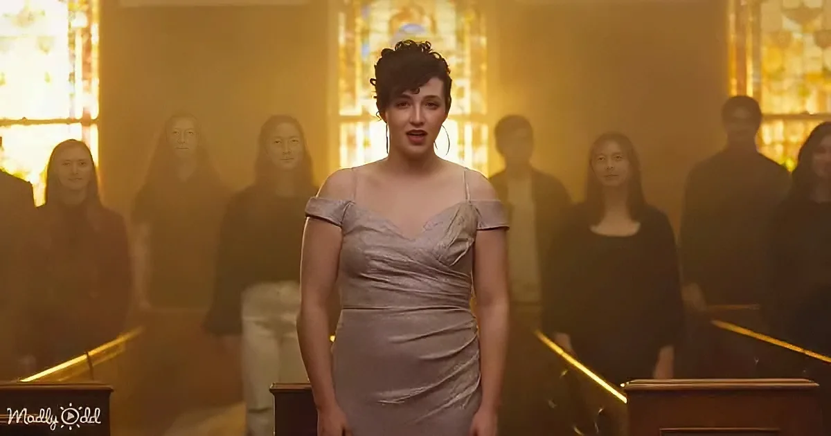 A woman in a light dress stands in a church, surrounded by a choir, with stained glass windows in the background.