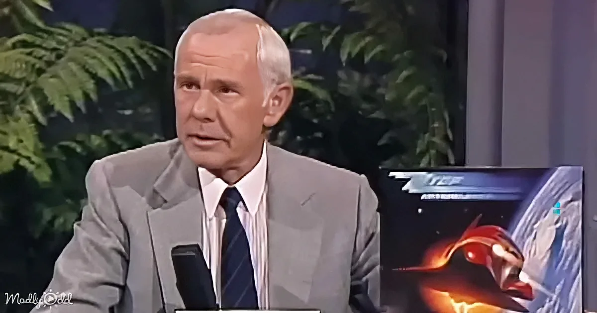 A man in a gray suit and tie is speaking, with a plant and a screen showing a red and yellow character in the background.