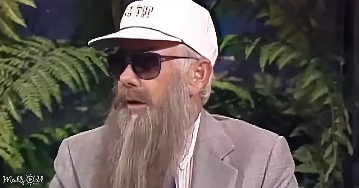 A man with a long white beard, wearing a white cap and sunglasses, stands in front of green foliage.