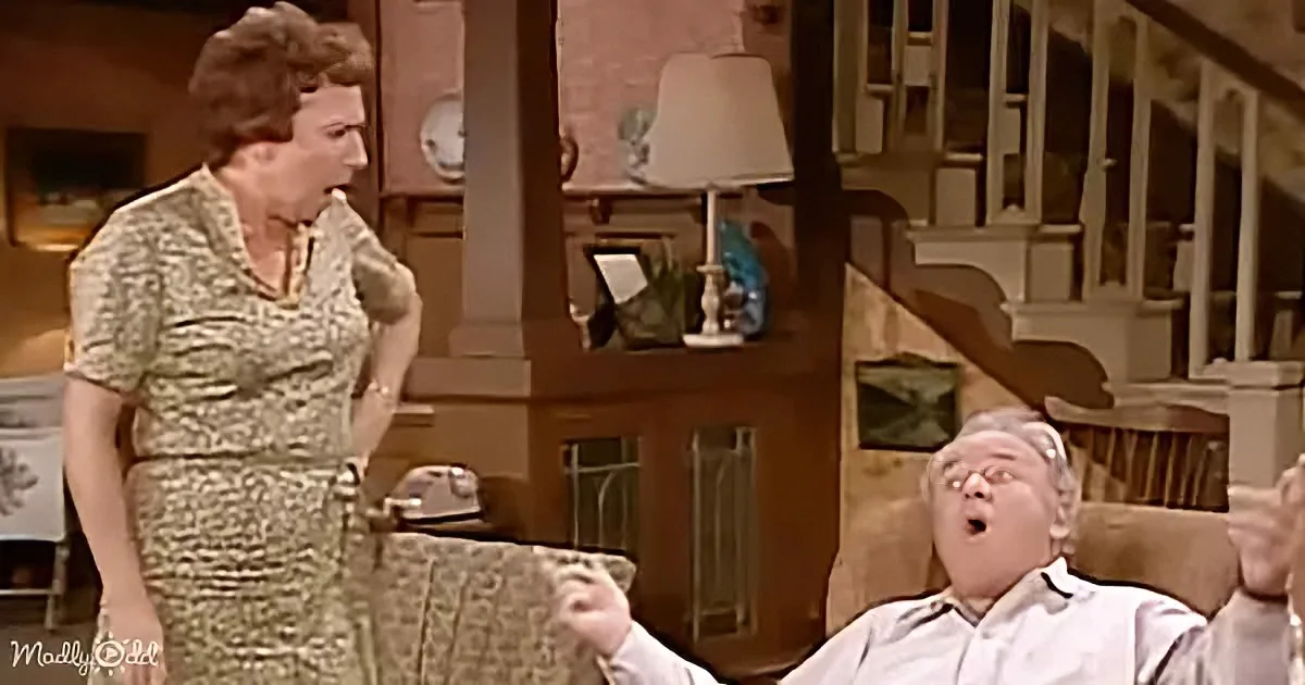 A woman in a patterned dress stands, gesturing, while a man in a white shirt sits in a chair, mouth open.