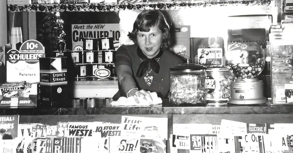 A woman stands behind a counter in a vintage store, surrounded by old advertisements and products.