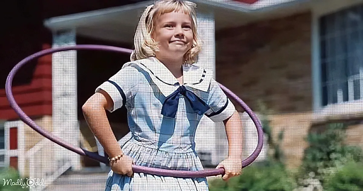 A girl in a sailor-style dress holds a hula hoop, standing in front of a house with her hands on her hips.
