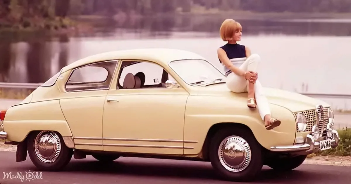 A woman sits on a cream-colored vintage car by a lake, wearing a sleeveless top and white pants.