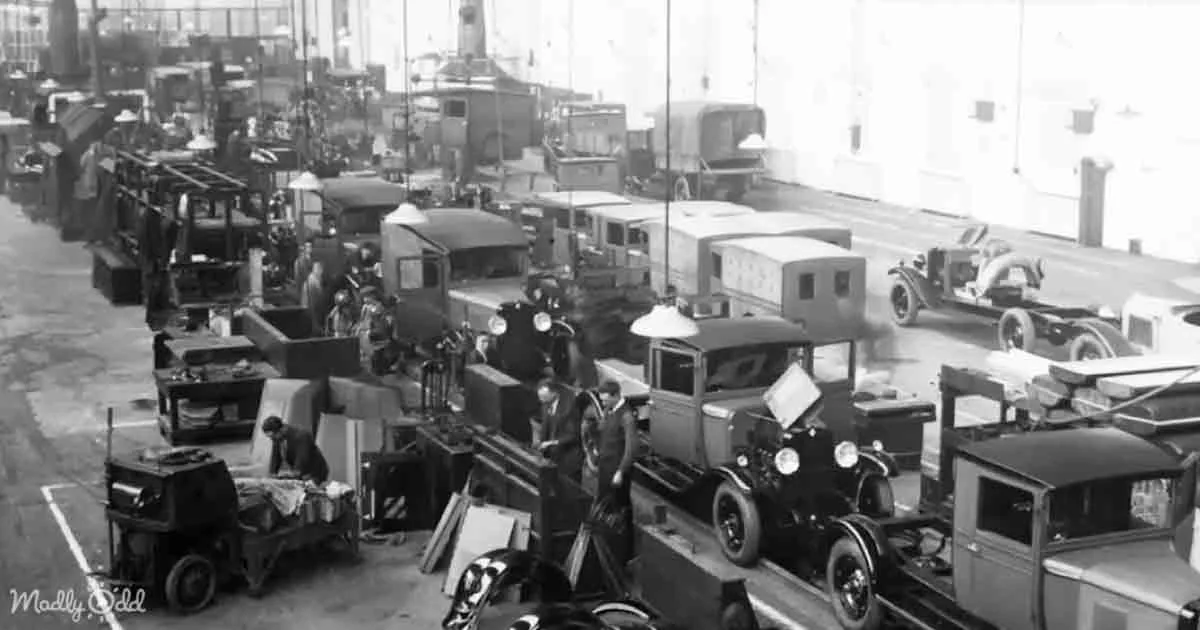 Workers assemble vintage cars on an assembly line in a bustling factory setting.