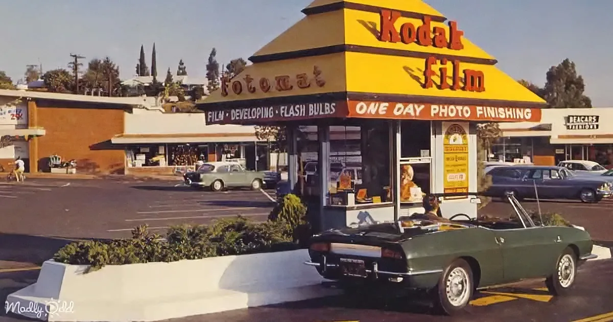 A Kodak Film Fotomat kiosk with a green car parked in front, surrounded by a parking lot and other buildings.
