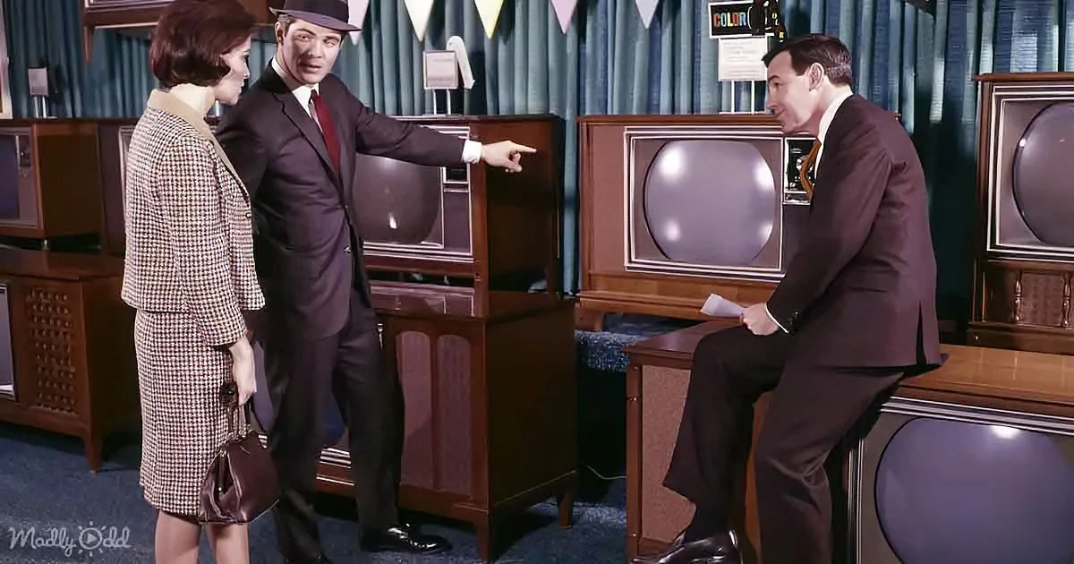 A man in a suit points at a television while a woman stands beside him, both in a showroom setting.