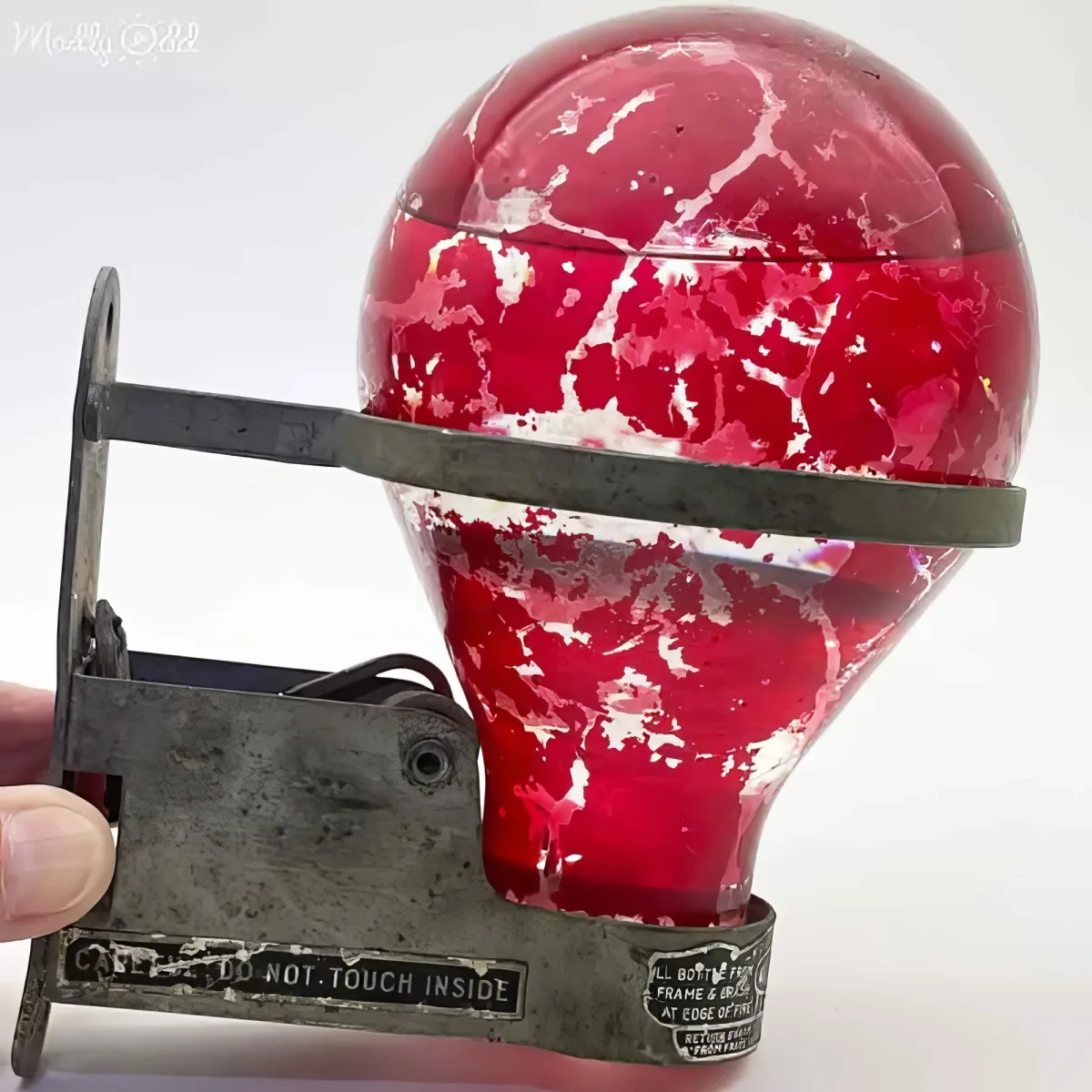 A red, cracked glass object is held in a metal clamp, with a hand visible on the left side.