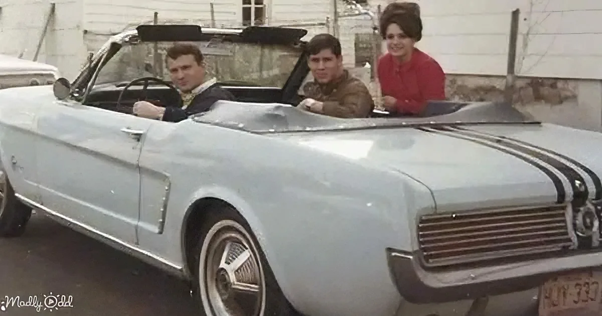 A vintage light blue convertible car with three people inside, parked in front of a house.