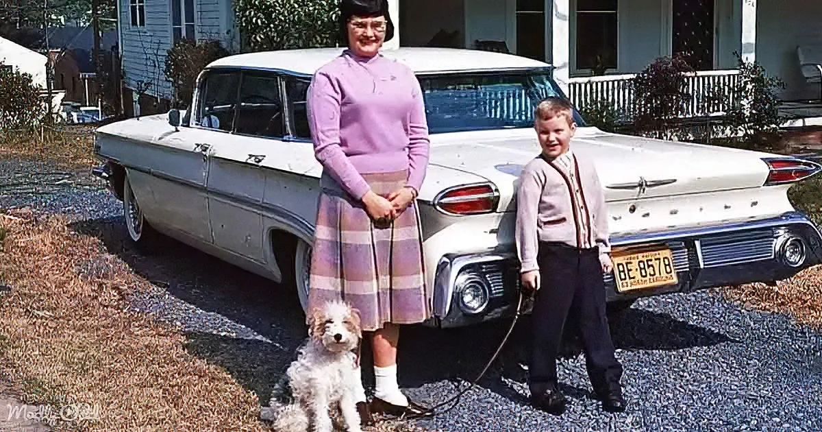 A woman and a boy stand with a dog in front of a classic white car, smiling.