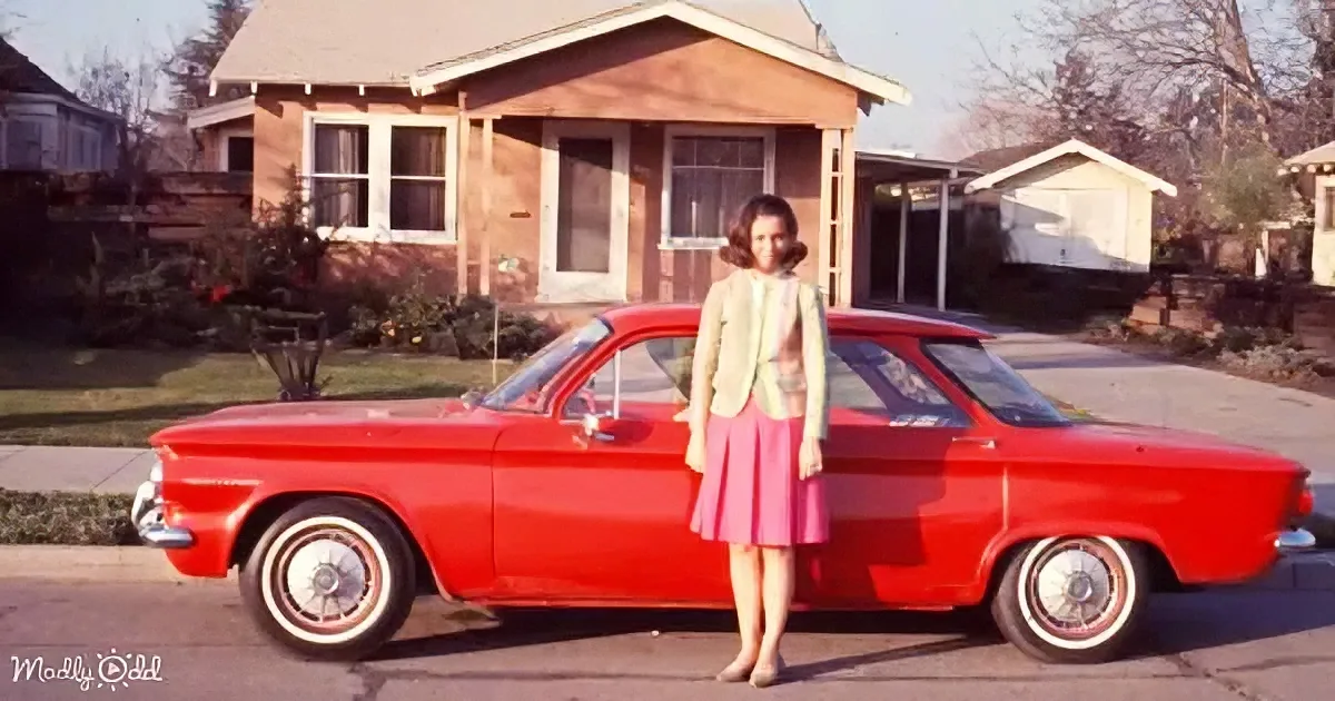 A woman in a pink skirt stands beside a bright red vintage car in front of a suburban house.