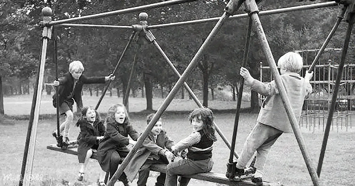Children playfully swing on a wooden playground structure in a park.