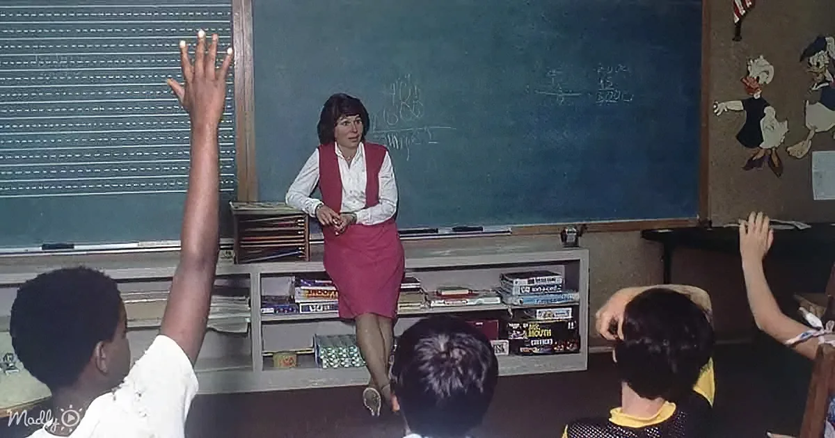A teacher stands at a chalkboard in a classroom, engaging with students raising their hands.