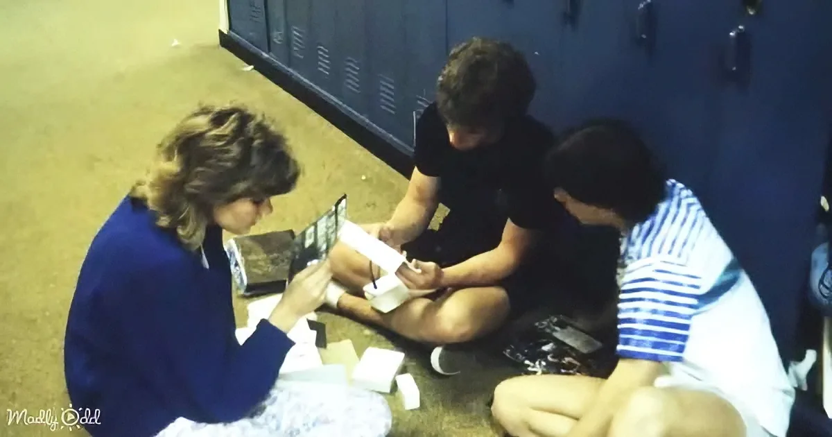 Three people sit on the floor near lockers, one holding a book, another a box, and the third looking down.