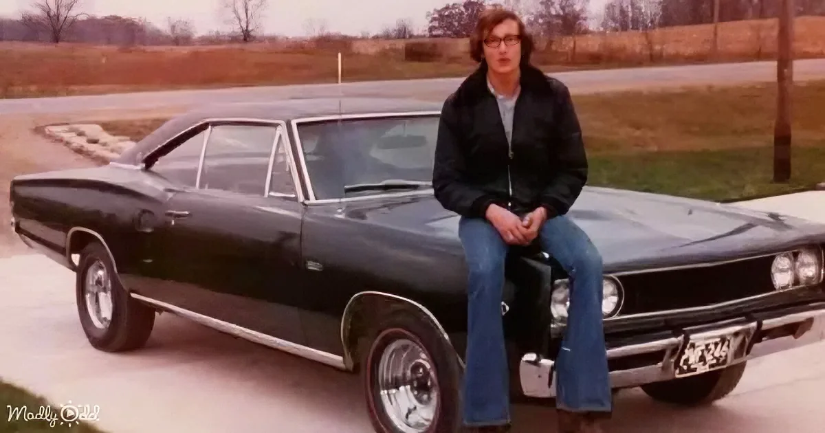 A man in a black jacket and jeans sits on a classic black muscle car, posing on a paved road with a field and trees in...