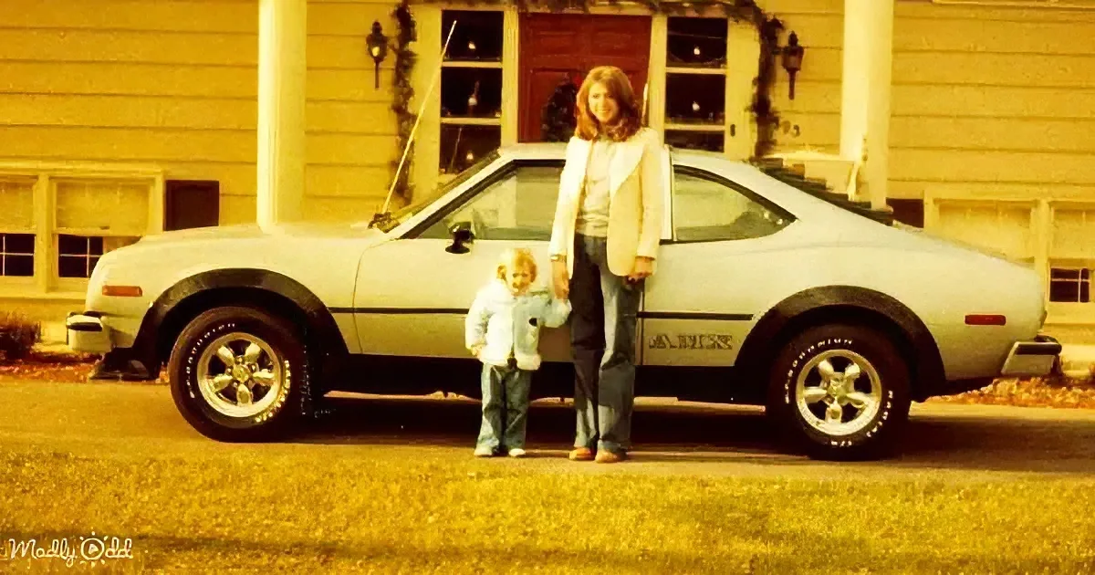 A woman and child stand beside a light-colored AMC car in front of a yellow house.